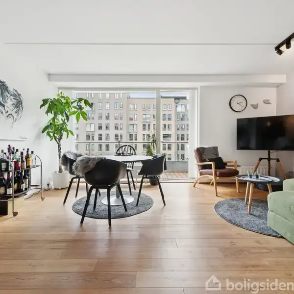 Stue med spisebord og stole i sort, grøn sofa og vægmonteret fjernsyn. Trægulv og vægkunst. Store vinduer med udsigt til bygninger. Planter og barvogn i hjørnet.