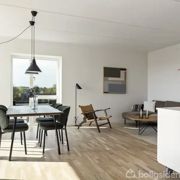 En spisestue med et bord og fem stole står under en sort loftslampe. Rummet har trægulv og hvidmalede vægge, og der ses en sofa samt et kunstværk i baggrunden.