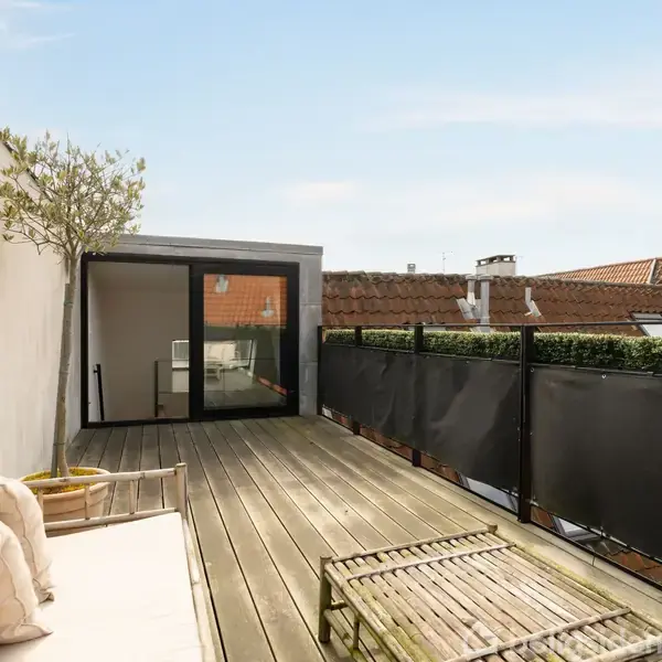 Terrasse med trædæk og sofa med puder, flankerende planter i krukker og et lavt bord. Glasskydedør åbner til et indendørs rum. Udsigt til tegltage og himmel.
