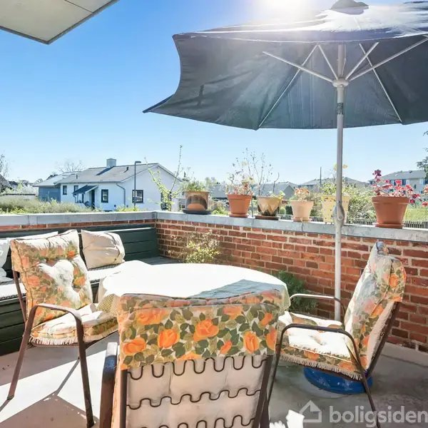 Terrassemøbler omkring et rundt bord med blomstrede hynder. Et stort parasol skygger området. Flere potteplanter står på en murstensvæg, og i baggrunden ses et hus og klar blå himmel.