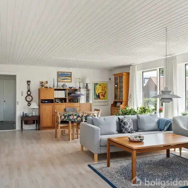 Stue med gråt sofaarrangement står omkring et trækaffebord på et tæppe. Rummet har hvidt loft, trægulv, og store vinduer med udsigt til en have.