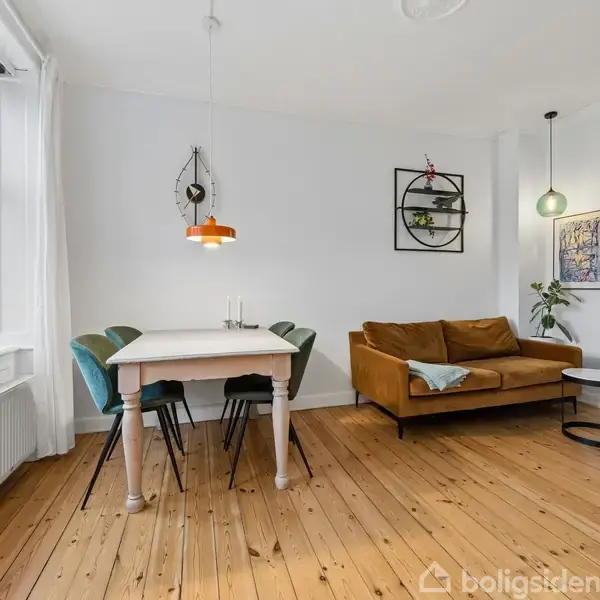 En hyggelig stue med trægulv indeholder et spisebord med fire stole og en brun sofa. Ved siden af er en bogreol og hængende lamper. Store vinduer giver naturligt lys.
