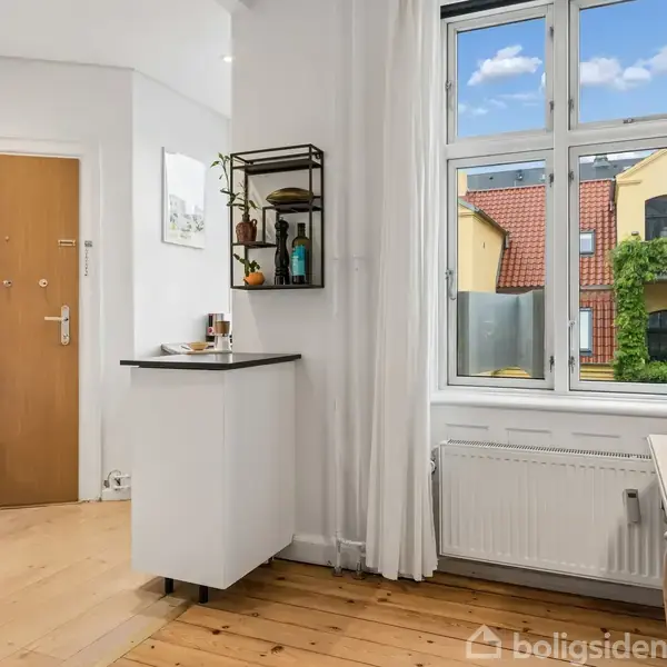 Et lyst køkken med trægulv, lille bord med sorte hylder, hvidt gardin ved stort vindue. Udsigt til røde tagsten og bygning med grønne slyngplanter.