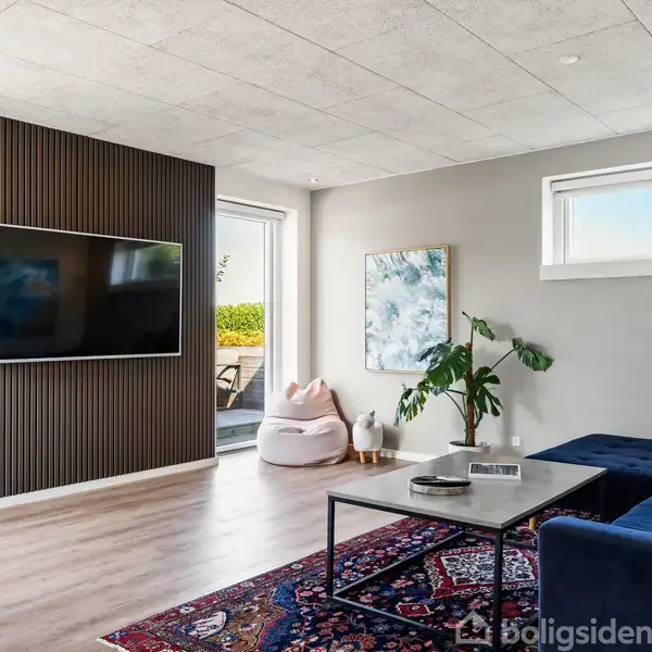Vægmonteret tv hænger på træbeklædt væg. Sofa står på farverigt tæppe i moderne stue med trægulv, store vinduer og planter. En dør fører ud til terrasse med udsigt til have.