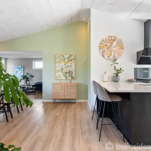 Et moderne køkken med en sort emhætte og bar, to højstole ved en hvid køkkenø, og grønne planter placeret omkring. I baggrunden ses en spisestue og opholdsstue.