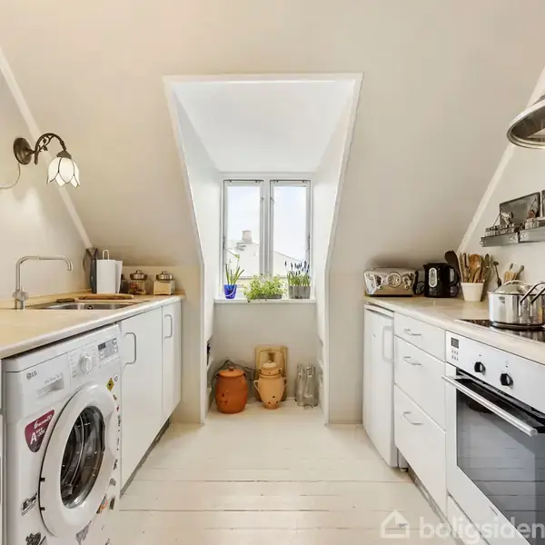 Hvidt, moderne køkken med vasketøjsmaskine og ovn, der står mod hver side af et skråloft. Et centralt vindue giver naturligt lys og udsigt til planter udenfor.