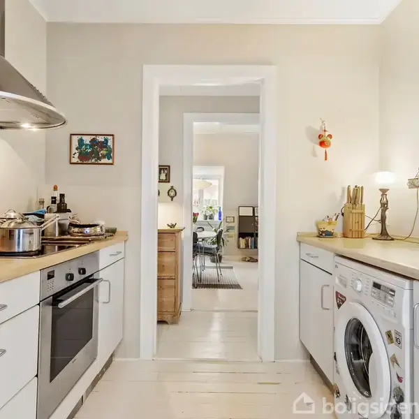 Et køkken med komfur og gryder til venstre, en vaskemaskine og lamperet på højre side. Der er en døråbning i midten, der fører til et lyst rum.