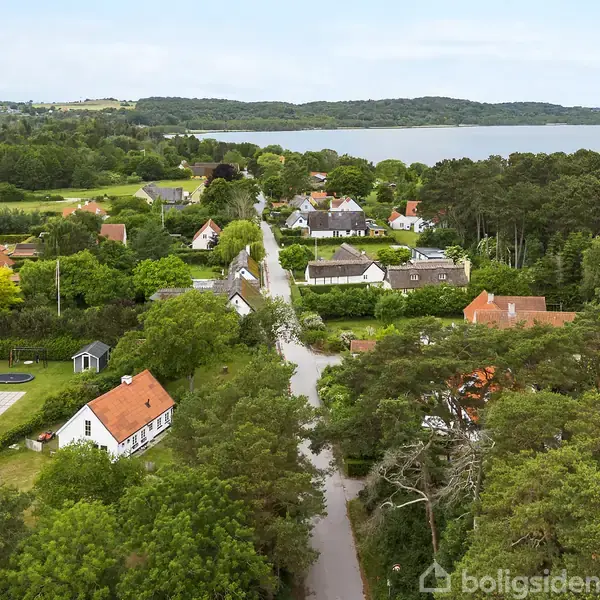 Landsbyhuse langs en snoet vej omgivet af tæt skov og marker. I baggrunden ses en sø med skovbevoksede bakker. Boligerne er spredt blandt frodig grøn vegetation.