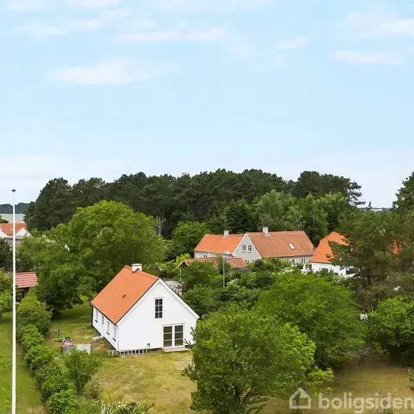Hus med rødbrunt tag står stille blandt frodige træer i et grønt, skovrigt boligområde. Flere lignende huse med røde tage er i nærheden, under blå himmel.
