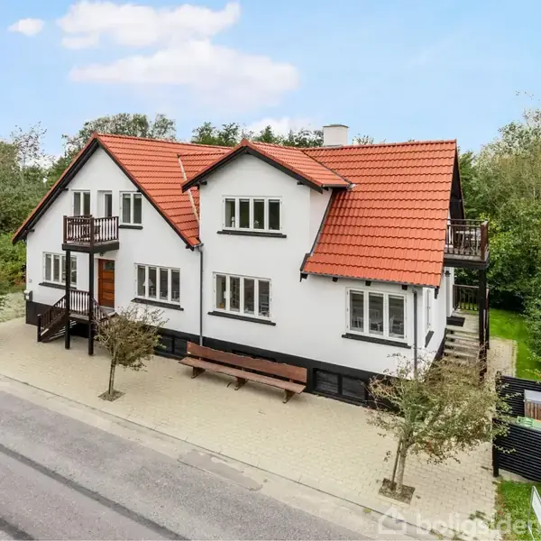 Hvidt hus med rødt tegltag og flere altaner. Det står på en hjørnegrund omkranset af grønne træer, med en asfaltvej foran.