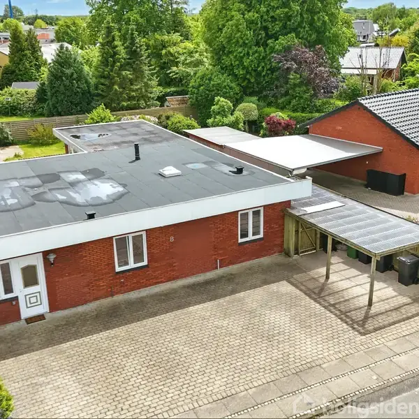 Rød murstensbungalow med fladt tag, står i et grønt, roligt boligområde. Foran huset er en indkørsel med carport.