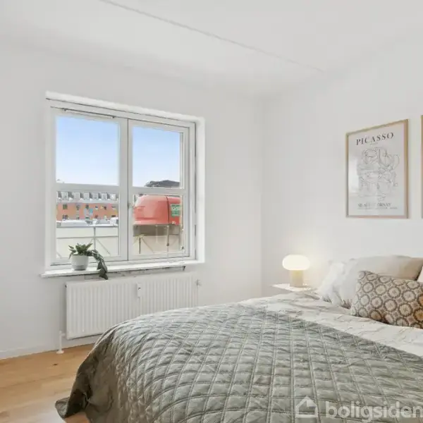 Soveværelse med seng dækket af grå sengetæppe og puder, ved siden af et lille bord med en lampe. Væggen har plakater med teksten "PICASSO" og "BOTANICA VERTIBI". Der er et vindue med udsigt.