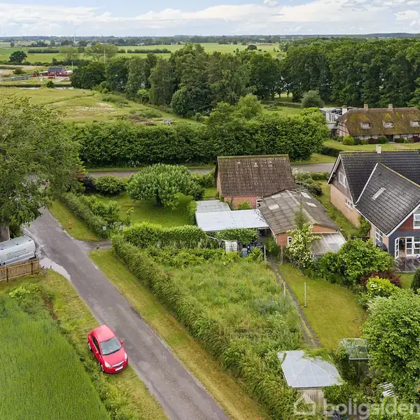 Huse står stille langs en vej omgivet af frodige marker og træer. En rød bil ses på vejen, mens en sø anes i baggrunden.