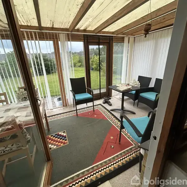 Et lukkede verandaområde med borde og stole med blå puder. Bordene har dekorationer, og gulvet har farverige tæpper. Store vinduer viser en grøn have udenfor.