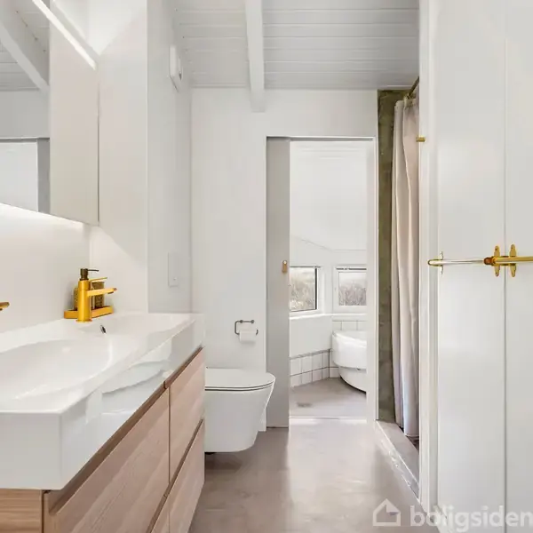 Dobbelthåndvask med gule vandhaner står i et lyst badeværelse med store spejle og træmøbler; til højre toilet og bruseområde med hvide vægge og gulv, det hele under hvidt loft.