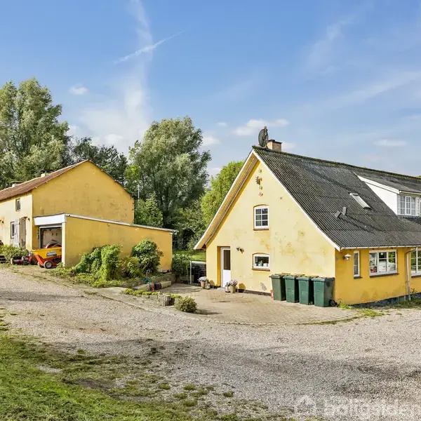 To gule bygninger står stille i et landligt område med grusvej og grønne omgivelser. Den ene bygning har skråt tag med vinduer, og der er affaldsbeholdere udenfor.