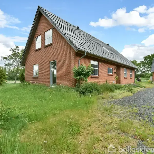 Rødt murstenshus med sort tag ligger på en grøn, delvist grusbelagt grund. Tre vinduer og en dør ses på siden. Solrig dag med få skyer på himlen.