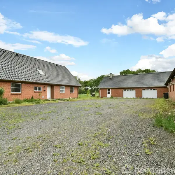 Rødt murstenshus med mørkt tag står stille ved siden af garager. Gruset gårdsplads omgiver bygningerne under blå himmel med skyer. Ingen tekst er i billedet.