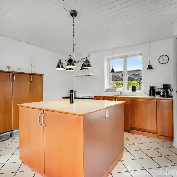 Et lyst køkken med hvidt loft og flisegulv. En stor træø med underskabe står centralt. Skabe i træ omgiver rummet. En dør fører ud, og en væg-ur viser tiden.