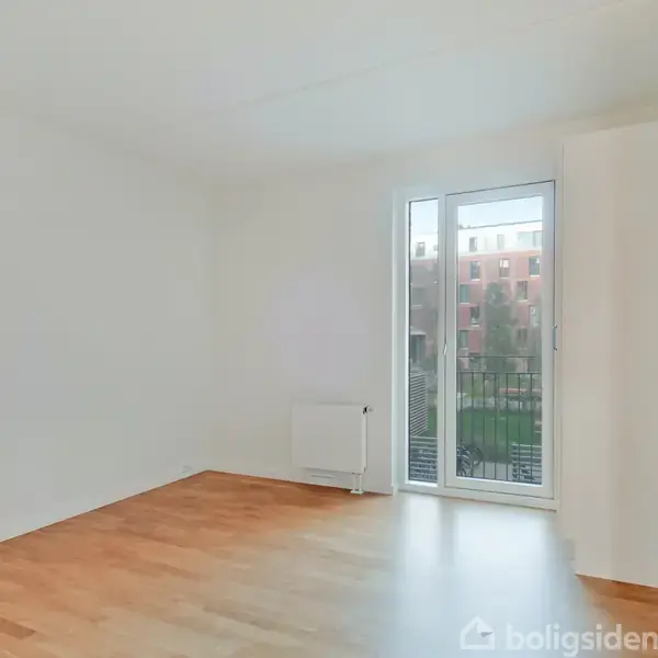 Et tomt rum med trægulv og hvide vægge, hvor en dør står åben mod en balkon. Udenfor ses en murstensbygning i baggrunden. Der er ingen tekst til stede.