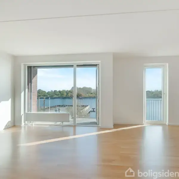 Stue med trægulv og store glaspartier, der åbner op til en udsigt over en sø og grønne områder udenfor. Lyset strømmer ind gennem vinduerne.