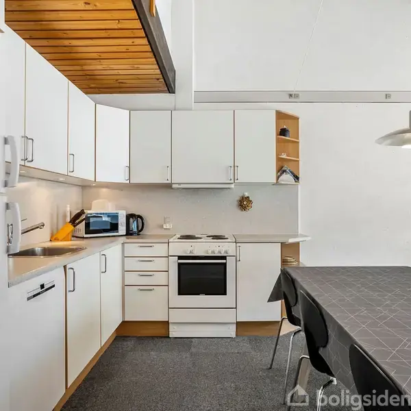 Køkken med hvidt skabsarrangement og komfur i hjørnet. En mikrobølgeovn står ved siden af vasken. Spisebord med mørk dug og stole i forgrunden. Loft af træ.