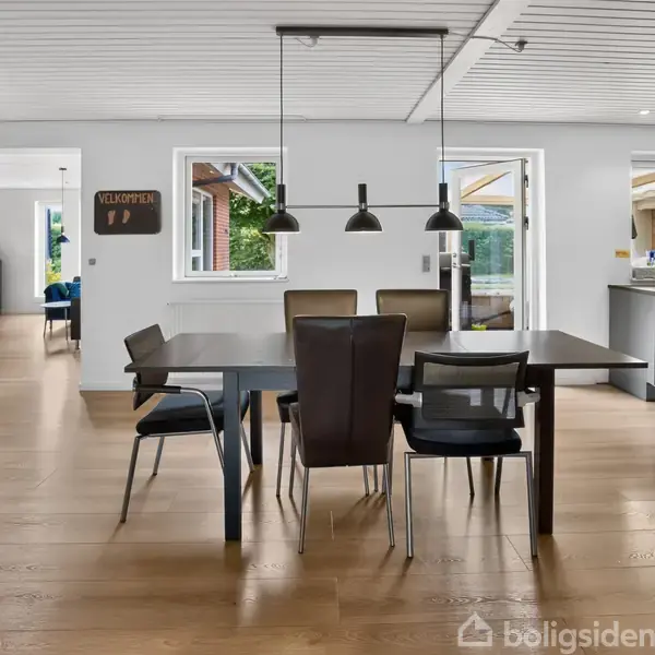 Spisebord med stole står i et køkken-alrum. Tre lamper hænger over bordet. Et skilt på væggen siger "VELKOMMEN". Store vinduer giver udsigt til en terrasse og have.