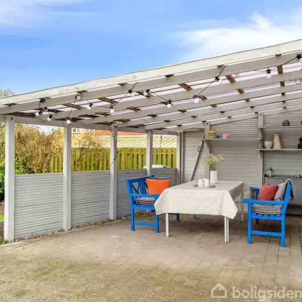 En overdækket terrasse med blå træmøbler, herunder en sofa og stol, omkring et bord med dug. Omkring er have og træhegn med lys guirlande under taget.