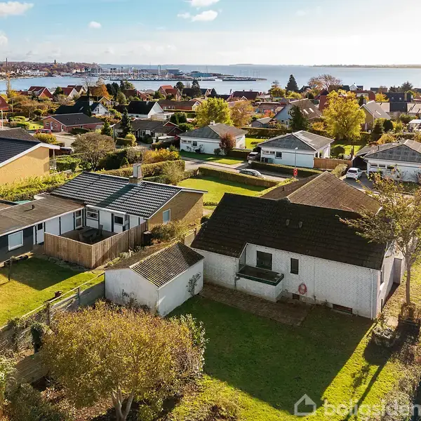 Flere enfamiliehuse med haver står stille i et villakvarter nær en kystlinje og havneområde. Farverige tage og efterårstræer omkranser området, med blå himmel og lette skyer i baggrunden.