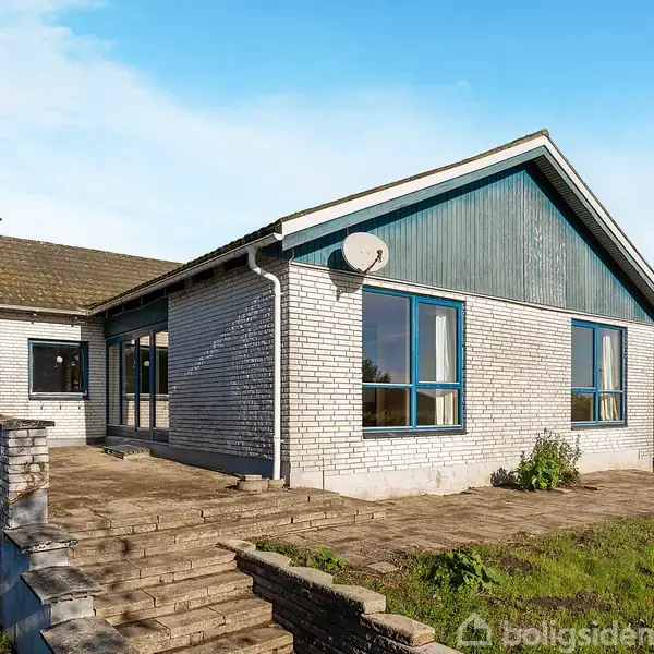 Et hus bygget af lyse mursten med blå detaljer står på en stenterrasse. Der er trapper, græsplæne og buske rundt om, og himlen er blå med få skyer.