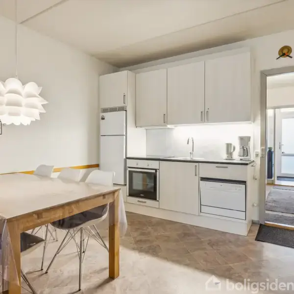 Køkkenbord med stole står under en hvid loftslampe. Køkkenet har hvide skabe og en vask, indlejret i en moderne indretning. En gang fører til flere rum.