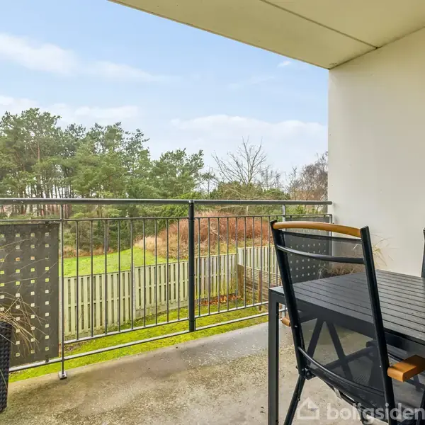 Et sort havebord med stole står på en balkon med udsigt til en grøn græsplæne omkranset af hegn og træer. En plantekrukke står ved rækværket.
