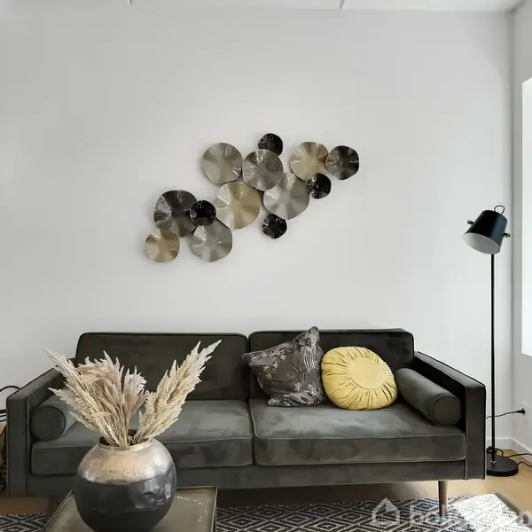 Grå sofa med puder står under dekorativ vægkunst. Ved siden af er en gulvlampe og stort vindue. Foran sofaen er et bord med vase og tørrede blomster.