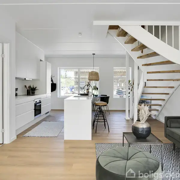 En moderne stue med spiraltrappe fører opad, en sofagruppe til højre og en køkken-ø i midten med tre barstole. Rummet er lyst og rummeligt.
