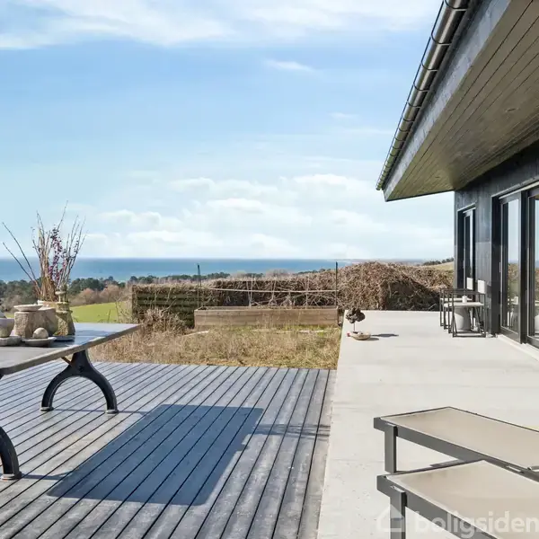Terrasse med sort træbord og stole vender mod havudsigt og åbne marker. Store glasdøre til højre reflekterer himlen, mens krukker dekorerer bordet.