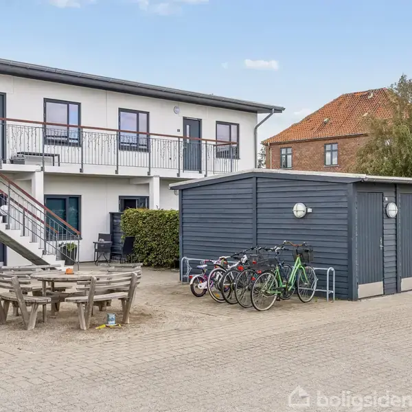 Cykler står parkeret foran en grå skurbygning med en trappe til venstre. Ovenpå trappen er en altan, og der er et træbord med bænke på gårdspladsen.