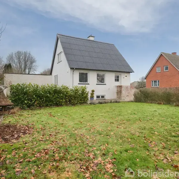 Et hvidt hus med sort tag står i en have med græs og buske. Et rødt hus er synligt til højre i baggrunden under en overskyet himmel.