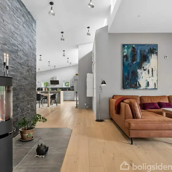 Brændeovn står ved stenvæg, med ild indeni. I stuen ses en brun sofa, dekoreret med en abstrakt maleri på væggen og en læselampe. Spiseområde i baggrunden.