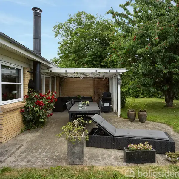 En moderne stue med en grå hjørnesofa og et sort sofabord, omgivet af kunst på væggene. Brændeovn brænder i hjørnet, adskilt af glasdøre med udsigt til terrasse.