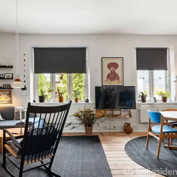Stue med sofa og gyngestol omkring et sofabord, i et lyst rum med billeder på væggene. Tv står mellem to vinduer med mørklægningsgardiner. Rundt spisebord med stole til højre.