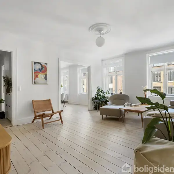 Stue med lyse trægulve, beige sofaer og grønne planter; sollys strømmer ind gennem store vinduer i en moderne, minimalistisk indretning. Maleri hænger på væggen.