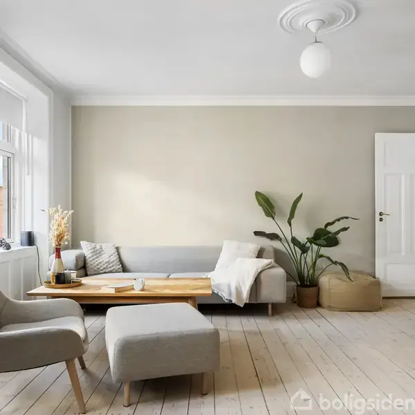 En stue med en grå sofa og lænestol arrangeret omkring et træbord. Plante og reol til højre. Værelset har lyst trægulv, hvide vægge og store vinduer mod venstre.
