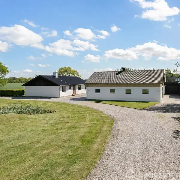 En hvid stuehusbygning med mørkt mønstret tag står stille på en velplejet græsplæne med grusindkørsel omgivet af træer. Skyer og blå himmel ses i baggrunden.