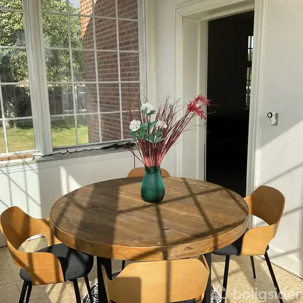 En rund træbord med en grøn vase og blomster i midten står i et rum med store vinduer. Sollys kaster skygger, og udenfor ses en grøn græsplæne.