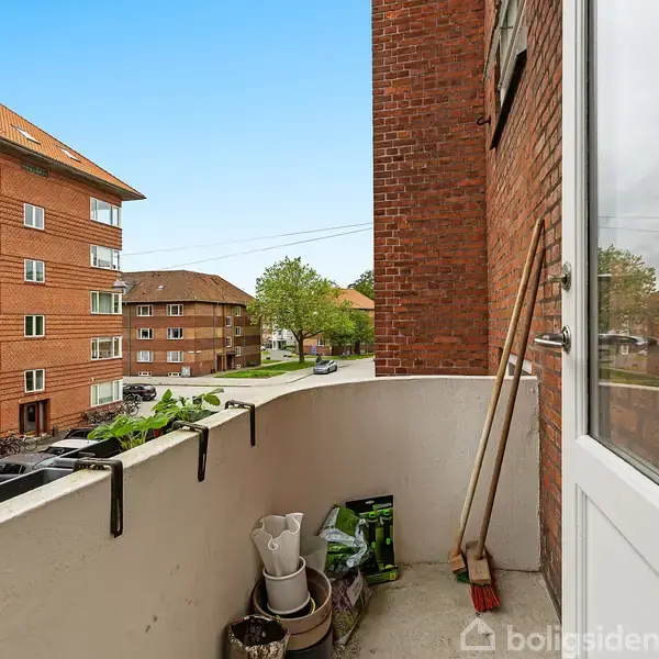 Balkon med potter og kost står stille; omgivet af røde murstensbygninger i et byområde.