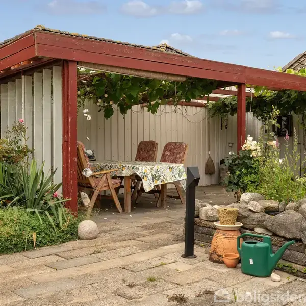 En overdækket terrasse med bord og stole; omgivet af planter og sten; beliggende ved siden af et hus med murstensvægge. En grøn vandkande og terracotta-potter står på terrassen.