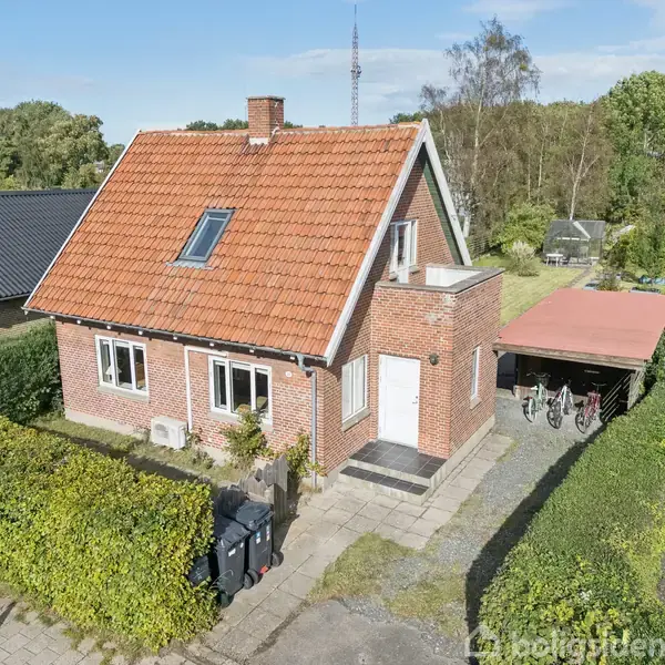 Rødt murstenshus med trekantet tag og skorsten, står stille i en frodig have. Ved siden af huset er en carport med cykler. Omkring er der træer og buske.
