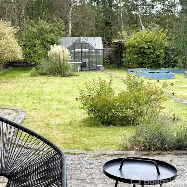 Et drivhus står stille i en velholdt have med grønne buske, græsplæne og et træbeklædt hegn omgivet af frodig vegetation.