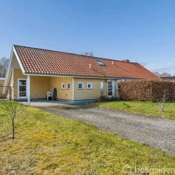 Et gult hus med rødt tegltag står i en have med flisebelagt indkørsel og omkransende hække. Træer ses i baggrunden under en blå himmel.