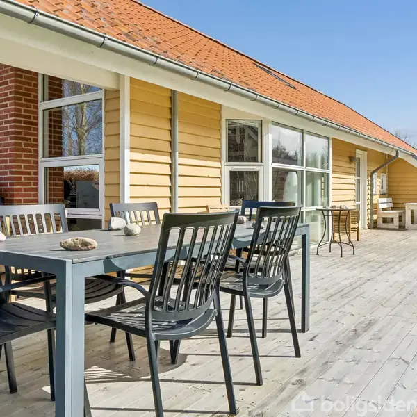 Udendørs terrasse med sort spisebord og stole står foran et gult træhus med rødt tag. Sollys skinner på området, omgivet af græsplæne.
